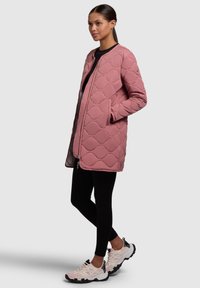 Veste matelassée rose avec un col rond, fermeture éclair à l'avant et poches latérales, associée à des leggings noirs et des baskets rose clair. Légère et texturée.
