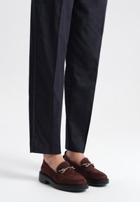 Brązowe zamszowe loafersy z detalem w postaci srebrnego �łańcuszka, zestawione z ciemnoniebieskimi dopasowanymi spodniami z białym przeszyciem wzdłuż szwów.