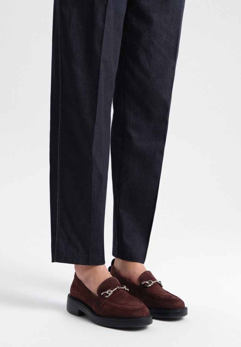 Brązowe zamszowe loafersy z detalem w postaci srebrnego łańcuszka, zestawione z ciemnoniebieskimi dopasowanymi spodniami z białym przeszyciem wzdłuż szwów.