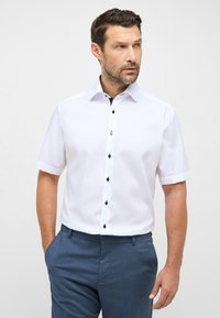 Camicetta bianca a maniche corte realizzata in tessuto morbido, con bottoni blu scuro e un dettaglio del colletto a contrasto. Abbinata a pantaloni blu scuro.