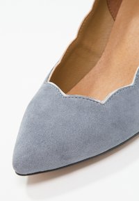 Chaussure plate en daim bleu clair à bout pointu avec bord en feston et doublure intérieure beige sur fond blanc.