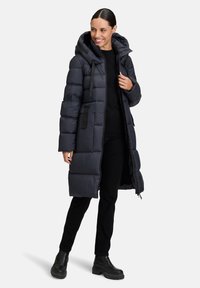 Cappotto imbottito lungo di un blu navy scuro con cappuccio, chiusura a zip, polsini elastici, tasche laterali e un design elegante trapuntato. Indossato con pantaloni neri e stivali.