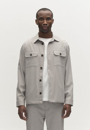 Férfi világosszürke zsebes felső ingben, fehér ing fölött, világosszürke nadrágban, sima háttér előtt áll.