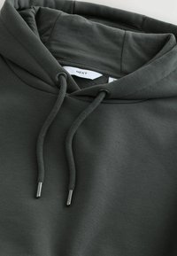 Tmavosivá mikina s hrubými šnúrkami a kovovými zakončeniami, zobrazujúca časť kapucne a značkový štítok "NEXT" vo vnútri goliera.