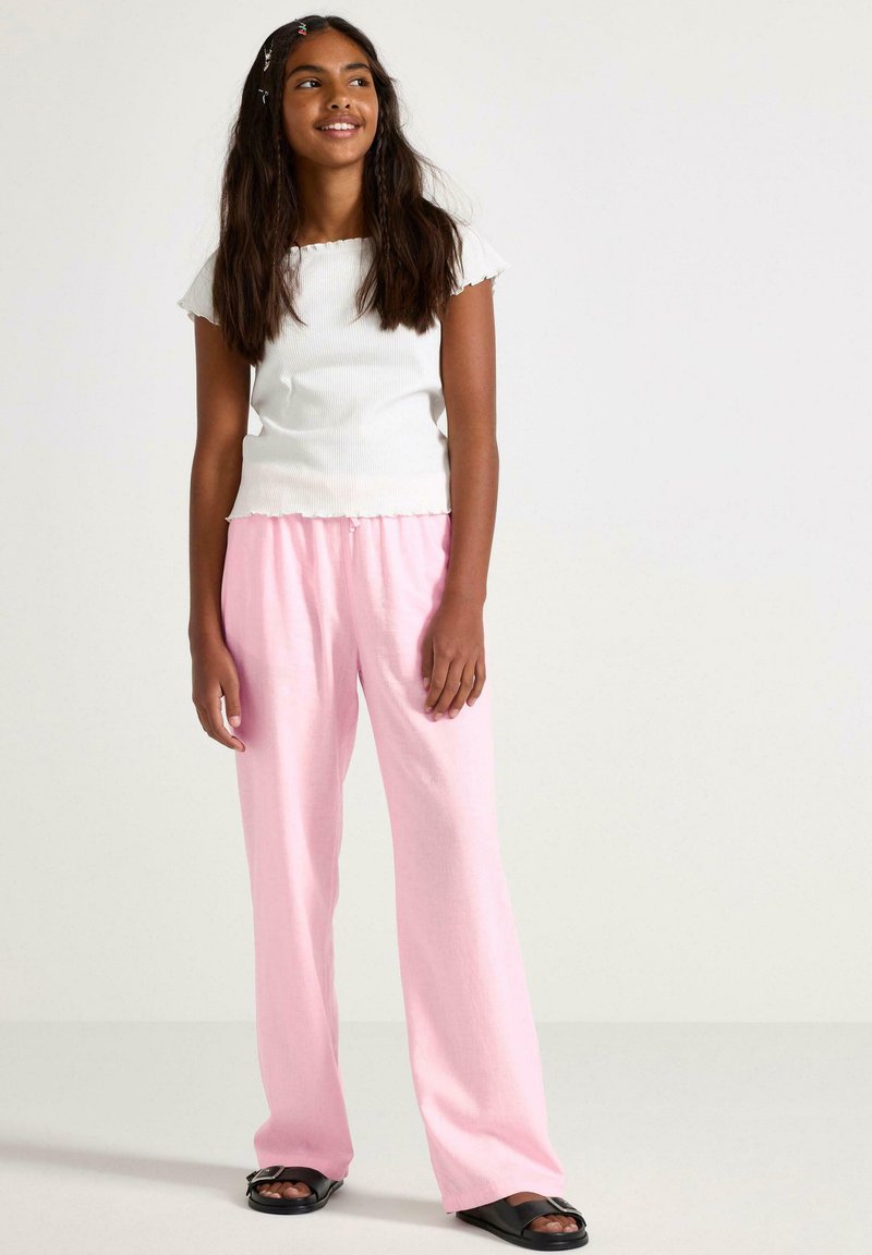 Witte geribde crop top met golvende randen, gecombineerd met roze loszittende broek. Model draagt zwarte slide-sandalen en staat tegen een effen achtergrond.