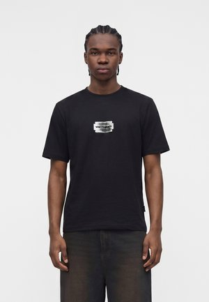 Junger Mann mit geflochtenem Haar steht nach vorne gewandt und trägt ein schwarzes T-Shirt mit einem kleinen weißen und grauen Grafiklogo auf der Brust.