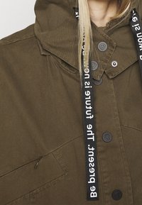 Olijfgroen hoodie met zwarte trekkoorden en witte tekst. Het heeft een knoopsluiting aan de voorkant en een zijzak met rits.