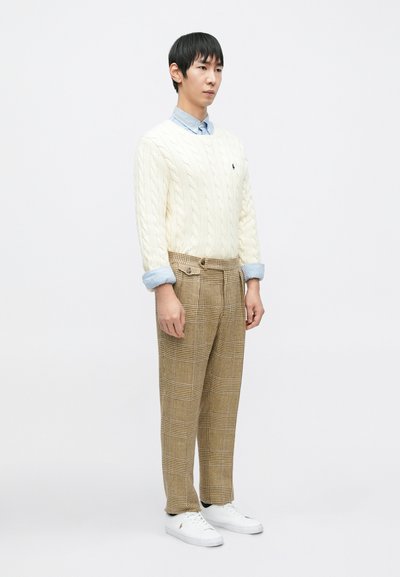 Camisola creme de malha com padrão trançado combinada com calças aos quadrados castanhas, com uma camisa azul clara com gola por baixo e ténis brancos.