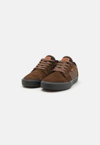Sapatilhas castanhas em camurça com solas de borracha pretas, ponta redonda, design com atacadores e etiqueta da marca laranja na lingueta, apresentando um perfil elegante.