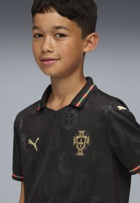 Črna nogometna majica z V-izrezom, z zlatim logotipom in poudarki, teksturiranim materialom ter ovratnikom z zelenimi, rdečimi in črnimi črtami.