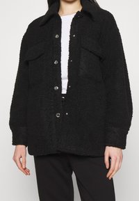 Svart fuskpälsjacka med krage, försedd med tryckknappar, två framfickor och en avslappnad passform. Mjuk textur med betydande värme.