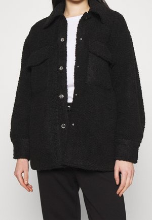 Mustat tekoturkistakki, jossa on kauluksellinen kaula-aukko, napitettu etuosa, kaksi etutaskua ja rento istuvuus. Pehmeä pinta ja huomattava lämpö.