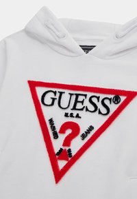 Biela mikina s kapucňou s výrazným trojuholníkovým logom v červenej a čiernej farbe, s textúrovanou výšivkou, ktorá znie "GUESS," "?" a "WASHED JEANS."
