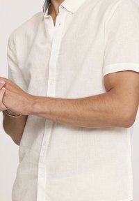 Chemise à manches courtes beige clair en lin avec un col boutonné. Présente un petit logo sur la poitrine et des accents de boutons blancs.