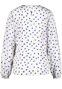 White blouse with blue polka dots, long sleeves, and a round neckline. Features a back keyhole opening and gathered cuffs.