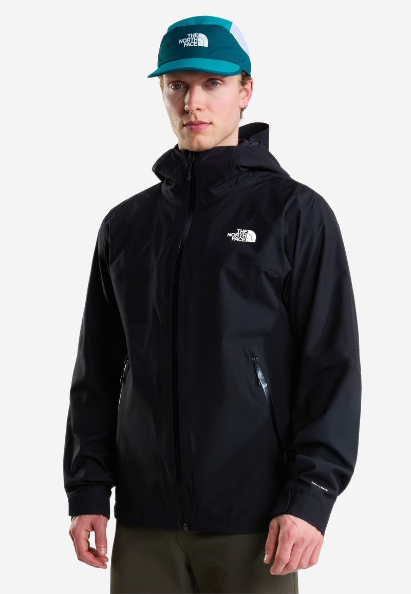 Νέος άνδρας με μαύρο μπουφάν North Face και πετρόλ πράσινο καπέλο North Face, στέκεται μπροστά σε ένα απλό λευκό φόντο.