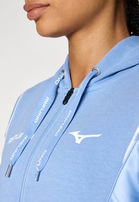 Sweatshirt azul claro com fecho éclair, material de algodão, logótipo branco em contraste, cordões ajustáveis e uma textura suave com um design justo.