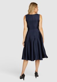 Marineblaues Etuikleid mit glatter Satin-Oberfläche, ärmellosem Design und Reißverschluss hinten. Der ausgestellte Rock reicht bis unter die Knie. Schwarze High Heels.
