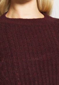 Gros plan sur une personne portant un pull en maille torsadée couleur bordeaux montrant les détails des motifs tricotés et la zone du cou.