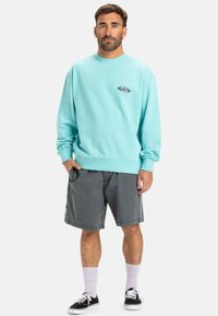 Light turquoise sweatshirt with ribbed cuffs, featuring a small logo on the chest. Paired with grey shorts and black sneakers.