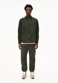 Man standing wearing dark green button-up shirt, matching cargo pants, beige sneakers, and light pink undershirt against plain light background.