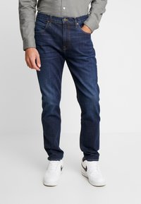 Granatowe dżinsy o prostym kroju, z pięcioma kieszeniami i zapięciem na guzik z przodu. W komplecie z białymi trampkami.