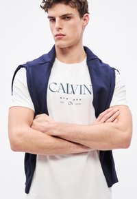 Suéter sin mangas de color azul marino sobre una camiseta blanca con un logo azul. El suéter tiene una textura suave y un ajuste cómodo, con un cuello redondo.