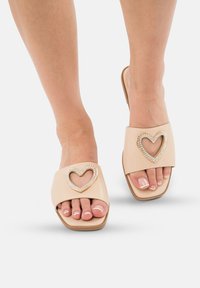Beige schuifsandalen hebben een brede band met een hartvormige uitsnijding, versierd met rhinestones. De zool is plat en vierkant van vorm.
