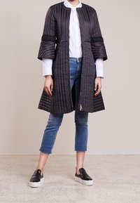 Svart quiltad kappa med rund halsringning och korta klockformade ärmar, kombinerad med blå korta jeans och svarta slip-on skor.