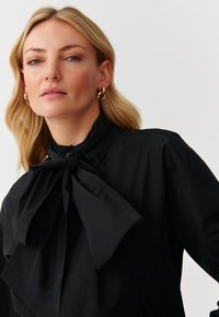 Blusa nera con colletto alto, dotata di un grande fiocco al collo. Tessuto liscio, maniche lunghe e pieghe sottili per un tocco di texture in più.