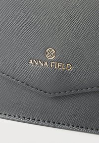 Κοντινή λήψη μαύρου δερματίνης με υφή, με χρυσό λογότυπο Anna Field και λεπτομέρειες ραφής στην επιφάνεια τσάντας ή πορτοφολιού.
