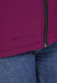 Lätt lila softshell-jacka med framdragkedja, svarta detaljer och broderad "Whistler"-logga vid fållen, kombinerad med blå jeans.