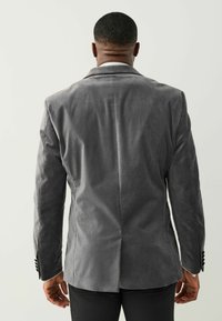 Grauer Samtblazer mit Reverskragen, einfacher Rückenventilation und knöpfbaren Manschetten. Glatte Textur mit tailliertem Schnitt, von hinten betrachtet.