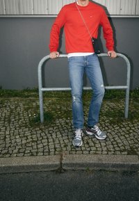 Sweatshirt rouge, chemise blanche en dessous, jean bleu clair déchiré, et baskets grises avec des accents rouges. Se tenant près d'une rambarde grise.