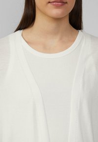 Weißer Cardigan mit lässiger Passform, aus weichem Material gefertigt. Hat einen runden Ausschnitt und dezente Nähte entlang der Kanten. Lange Ärmel sind sichtbar.