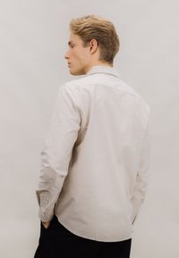 Lys beige langermet skjorte med klassisk krage, knappede mansjetter og en glatt bomullstekstur. Brukt sammen med svarte bukser, vist bakfra.