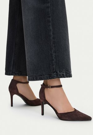 Mujer con zapatos de tacón alto de ante marrón oscuro cerrados en la punta con tiras en el tobillo, combinados con vaqueros negros de pierna ancha sobre un fondo blanco.