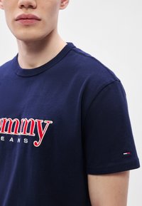 Camiseta de algodón azul marino con logo "Tommy" bordado en rojo y blanco, mangas cortas y un sutil detalle del logo en la manga.