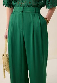 Groene, geplooide broek van gladde stof, met een hoge taille en zakken, gecombineerd met een textuurtas in goudkleur.