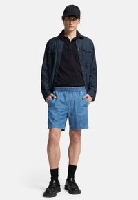 Camicia di denim con due tasche sul petto, polo nera, pantaloni corti leggeri con vita elastica blu chiaro, calze nere e scarpe nere. Completo semplice e casual.