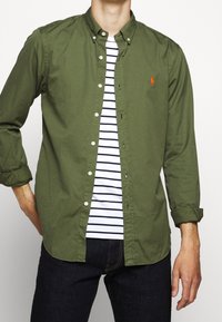 Olivgrön skjorta med knappar ovanpå en vit och marinrandig t-shirt, med en liten röd logotyp, i kombination med mörka jeans.