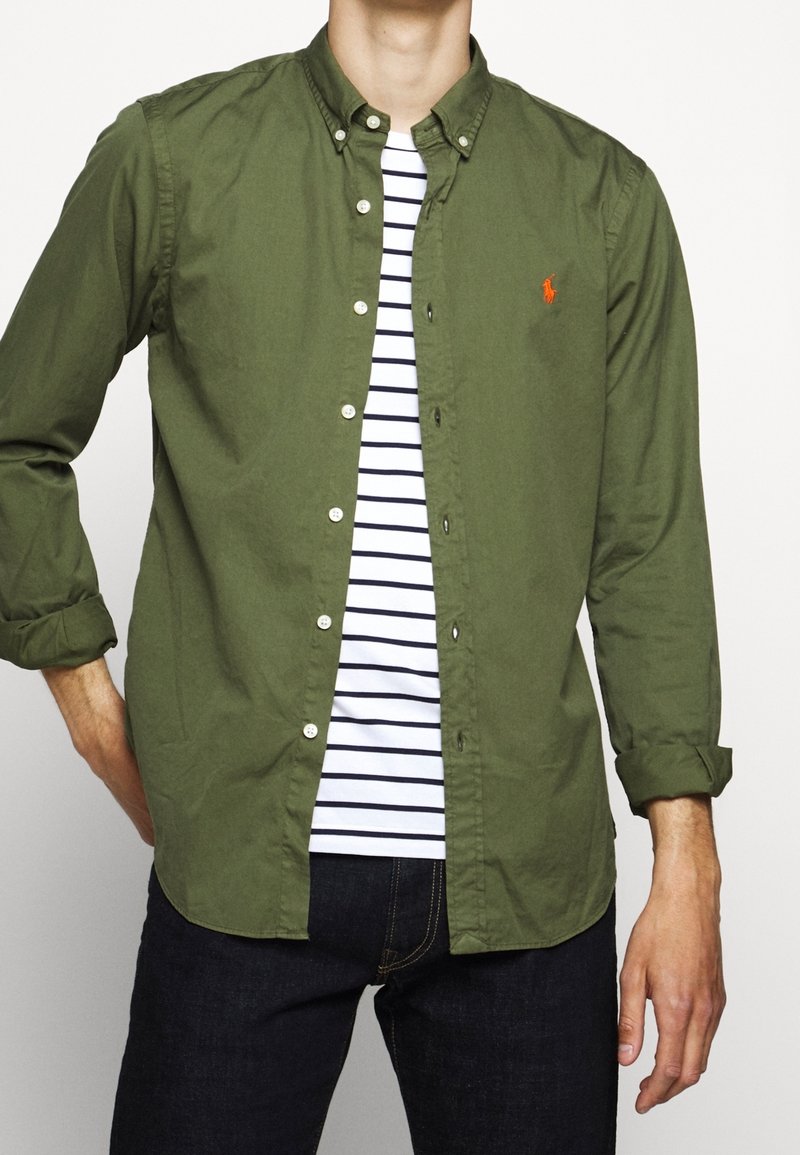 Olivgrön skjorta med knappar ovanpå en vit och marinrandig t-shirt, med en liten röd logotyp, i kombination med mörka jeans.