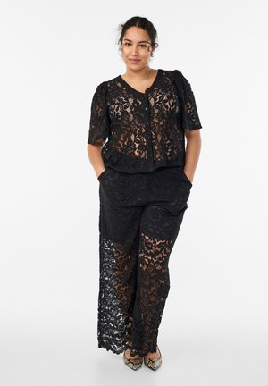 Frau trägt ein schwarzes, kurzärmeliges Spitzentop mit Blumenmuster und dazu passende, weit geschnittene Hosen mit spitzen Schuhen mit Schlangenhautmuster, steht mit den Händen in den Taschen.
