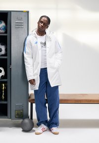 White long coat with blue stripes, gray graphic t-shirt, blue jeans, and red-accented sneakers. Locker and sports equipment in background.