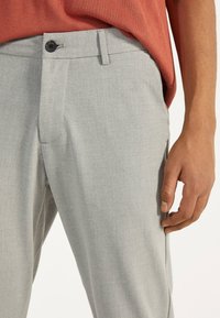 Ljusgrå byxor i ett strukturerat tyg, med knappstängning och slät framsida. Sidofickor synliga på varje sida.