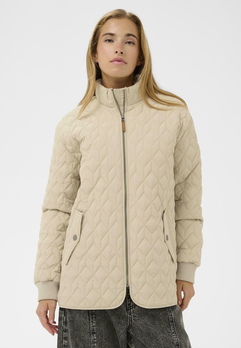 Veste matelassée beige avec un col montant, fermeture éclair sur le devant et poches latérales. Dispose de poignets côtelés et d'une texture lisse et douce.