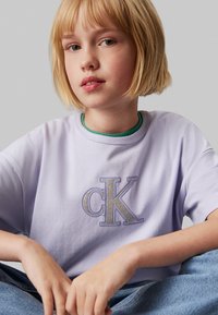 Camiseta de algodón de color púrpura claro con un logo "CK" bordado en plata en el frontal, escote redondo y corte relajado. Llevada sobre una blusa verde.