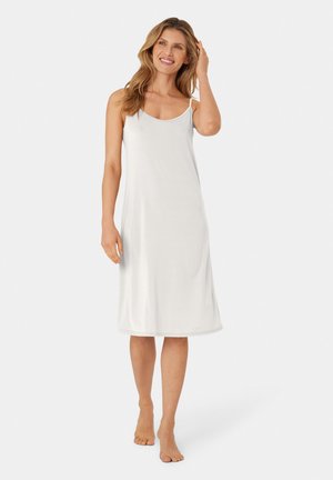 Frau lächelt, steht barfuß, trägt ein ärmelloses, weißes, knielanges Unterkleid und berührt mit einer Hand ihr Haar vor weißem Hintergrund.