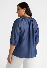 Blusa in chambray blu con maniche a tre quarti, scollatura arricciata e dettagli plissettati sulle maniche. Tessuto morbido, vestibilità casual.