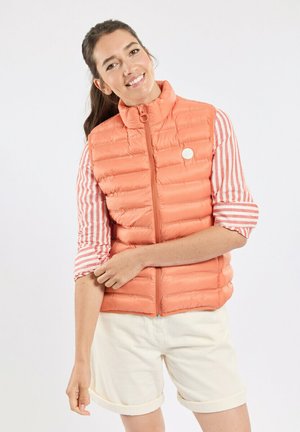 Femme souriante aux cheveux foncés portant un gilet matelassé orange par-dessus une chemise rayée retroussée et un short crème sur fond blanc.